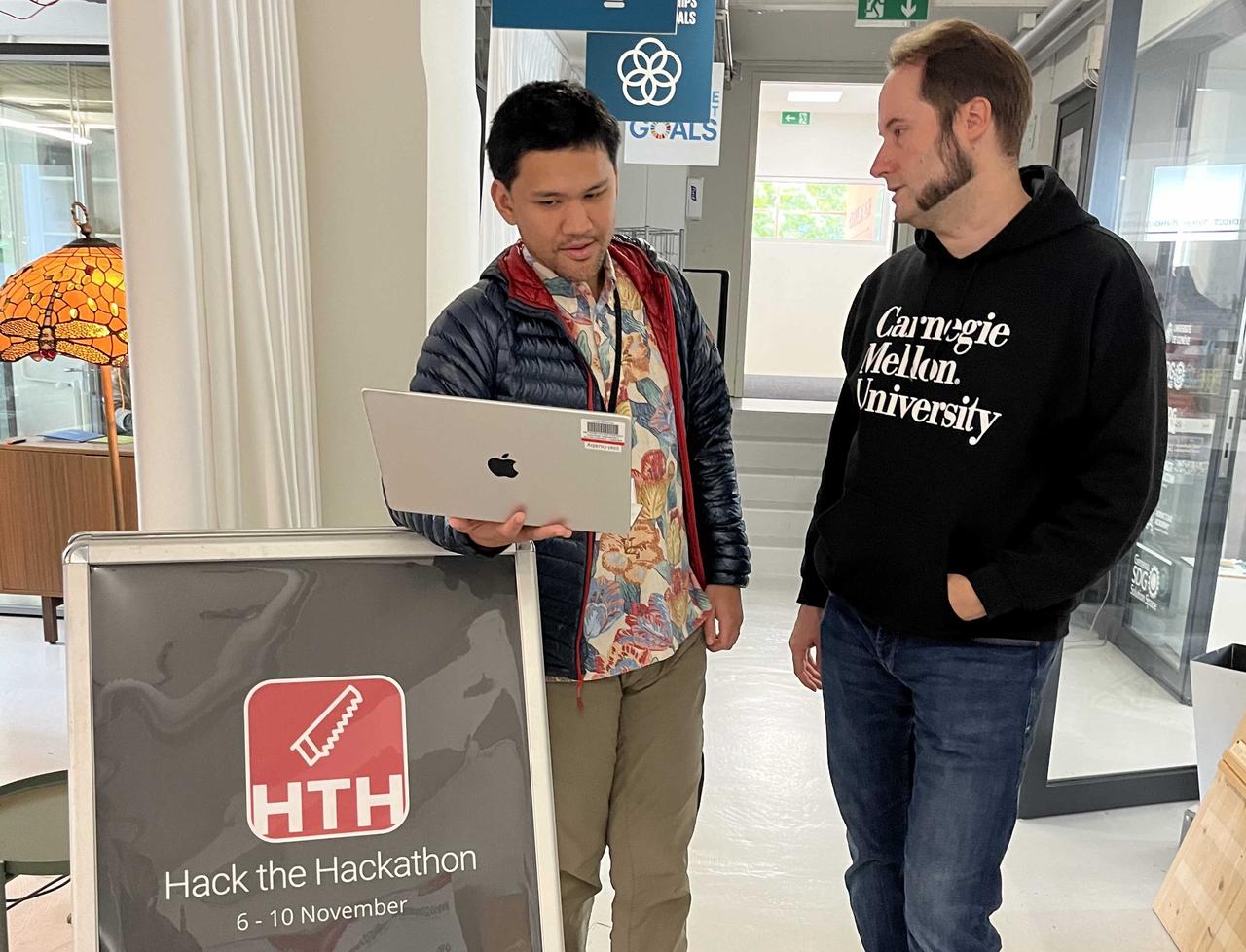 Two men standing next to a sign saying 'hack the hackathon', one holding a computer. They are in deep discussion.