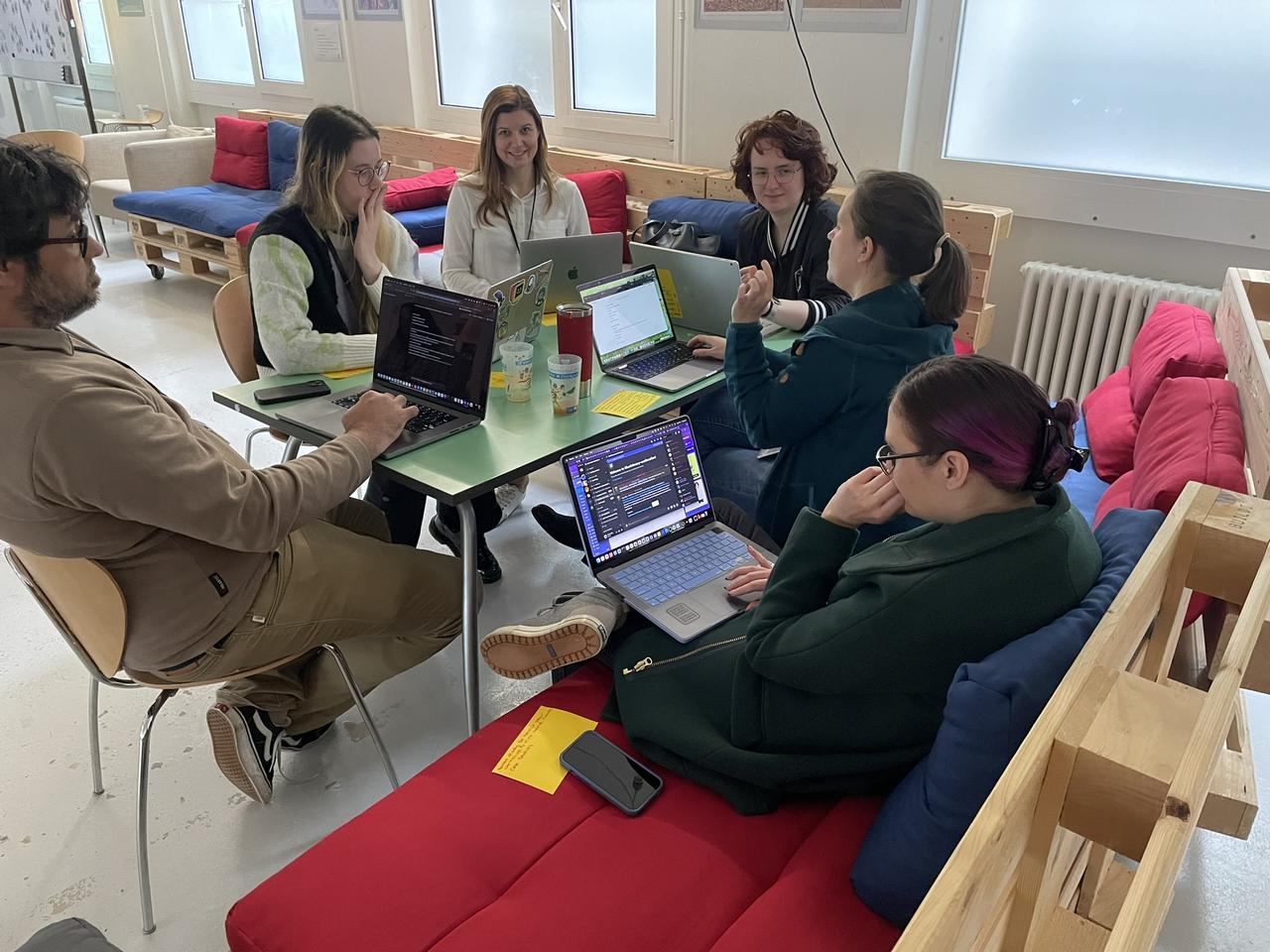 A group of people in a huge hangar turned into a Makerspace, sitting around tables with laptops and listening to someone at the front give a presentation. In the back, there is a big red London bus that's part of the furniture.